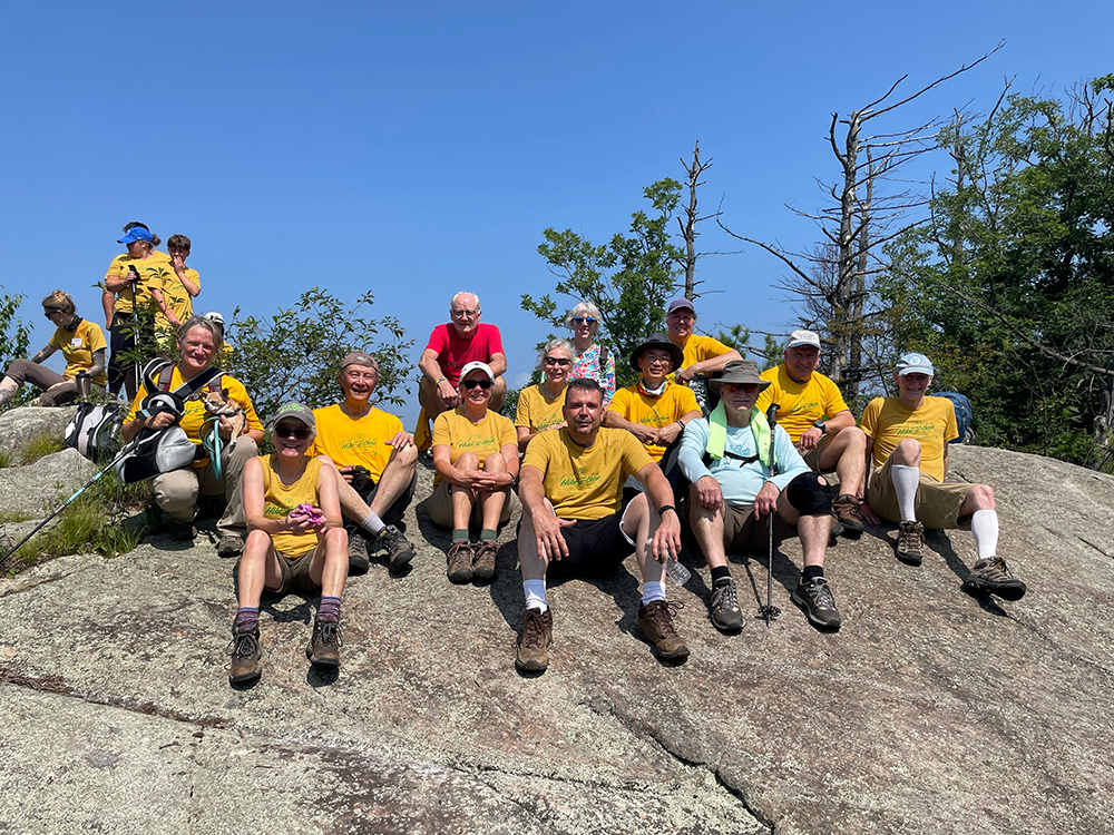 A dozen people wearing bright yellow tshirts sit facing the camera on a large exposed rock at a mountain summit. Open blue sky is behind them.